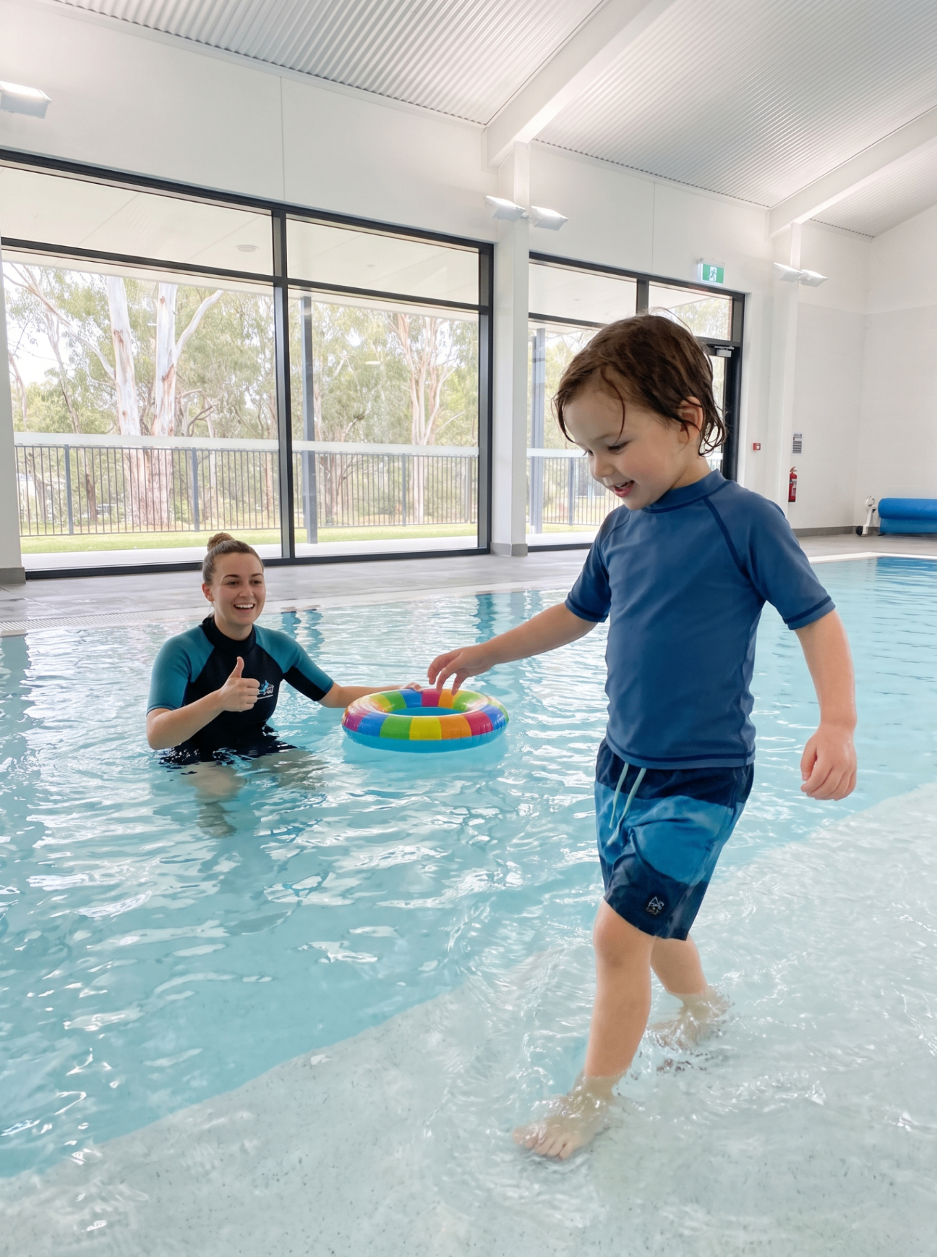 A young child playing at the edge of an indoor pool while a swim instructor smiles and supports them from the water